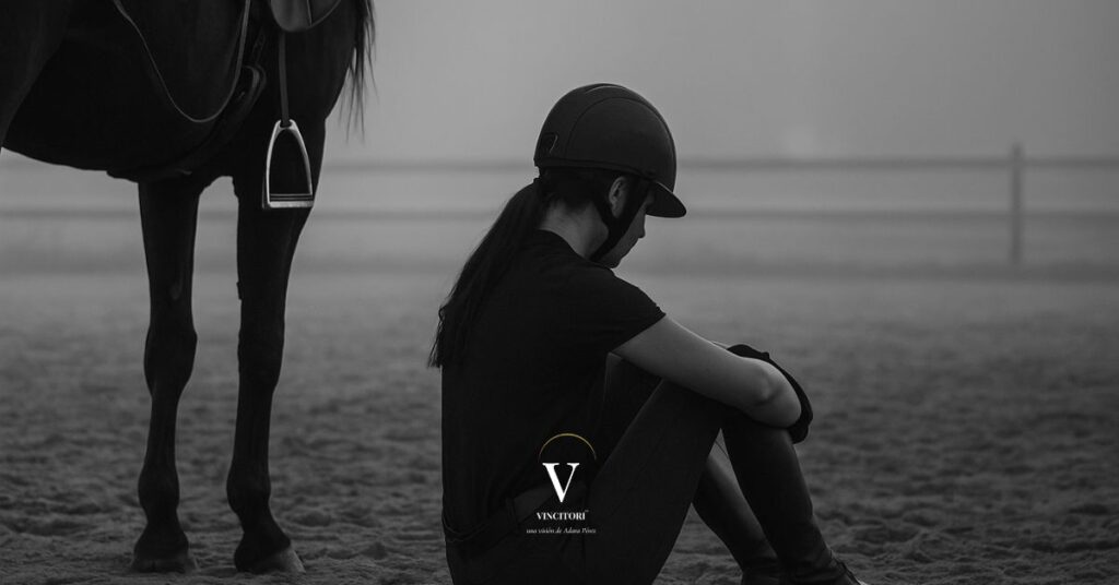Amazona sentada junto a su caballo, en blanco y negro, con el cuerpo agotado y la cabeza gacha; imagen que representa el cansancio emocional y la lucha silenciosa. Vincitori Adara Pérez